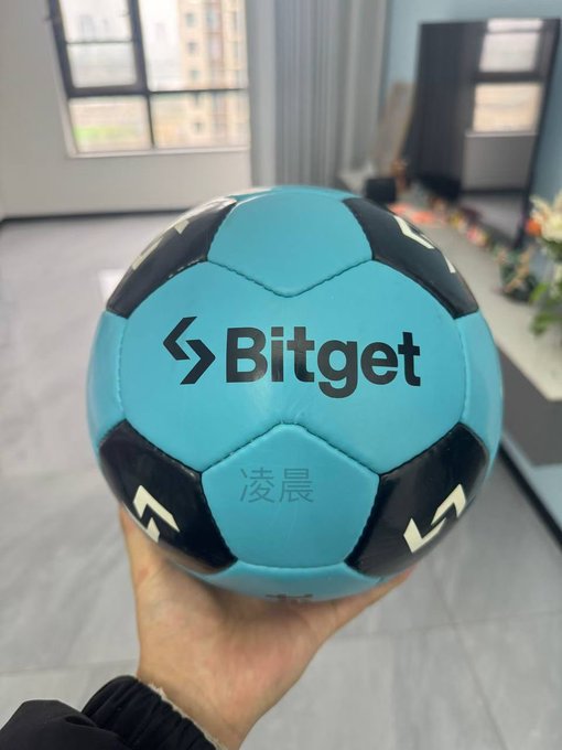 A hand holds up a light blue and black soccer ball prominently featuring the 'Bitget' logo and Chinese characters '凌晨', with an office interior visible in the background.