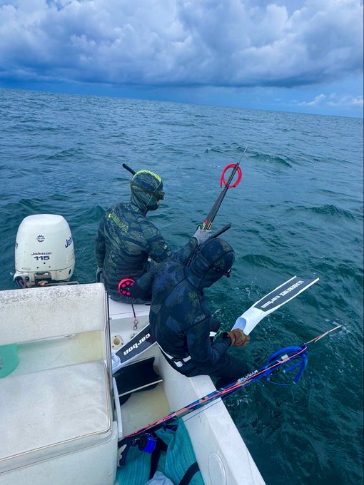 Two spearfishers in camouflage wetsuits sit on the edge of a boat on the open ocean, one holding a long speargun with a red reel, alongside a Johnson 115 outboard motor.