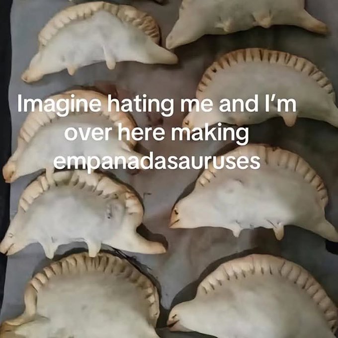 A tray of unbaked empanadas shaped like dinosaurs (stegosauruses) with crimped edges, overlaid with the text 'Imagine hating me and I'm over here making empanadasauruses'.