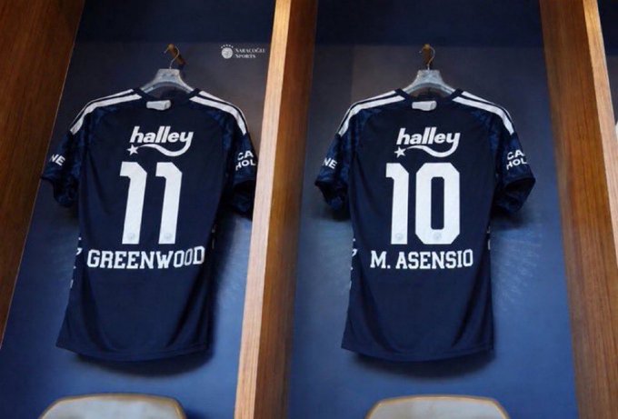 Two dark blue Fenerbahçe jerseys hang side by side in wooden lockers, displaying the names "GREENWOOD" (#11) and "M. ASENSIO" (#10) with the "halley" sponsor logo. The image represents a new era for the club, fitting the post's "AURA." theme by showcasing fresh, high-profile signings ready to make an impact.