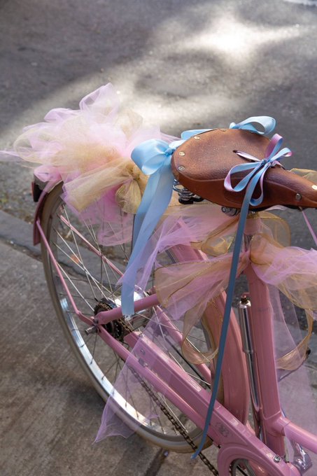 The rear section of a pink bicycle decorated with colorful tulle and blue ribbons, featuring a brown leather seat.