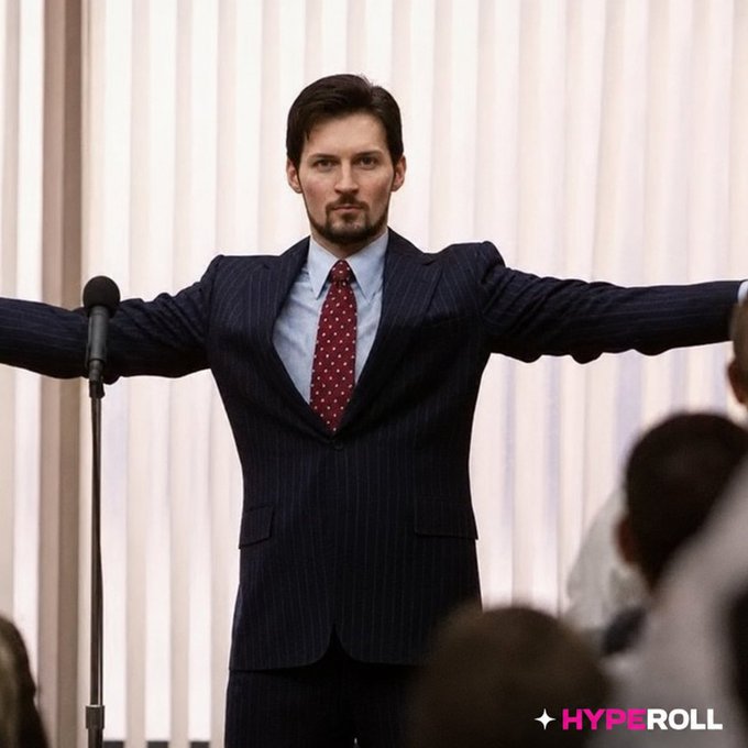 A man in a navy pinstripe suit and red polka-dot tie stands with arms outstretched at a podium microphone, facing a crowd, with the "HYPEROLL" logo in the corner—suggesting he's presenting or hyping up an audience, mirroring the post's promotional giveaway pitch.

Wait—looking again, the man's pose with arms wide open evokes a welcoming or grand gesture, fitting the "DROP your telegram handle" call-to-action vibe, while the "HYPEROLL" branding ties directly to the post's promotional giveaway content.

A suited man with arms outstretched at a podium projects charisma and command, visually amplifying the post's energetic crypto giveaway hype, with the "HYPEROLL" logo anchoring the promotional brand.