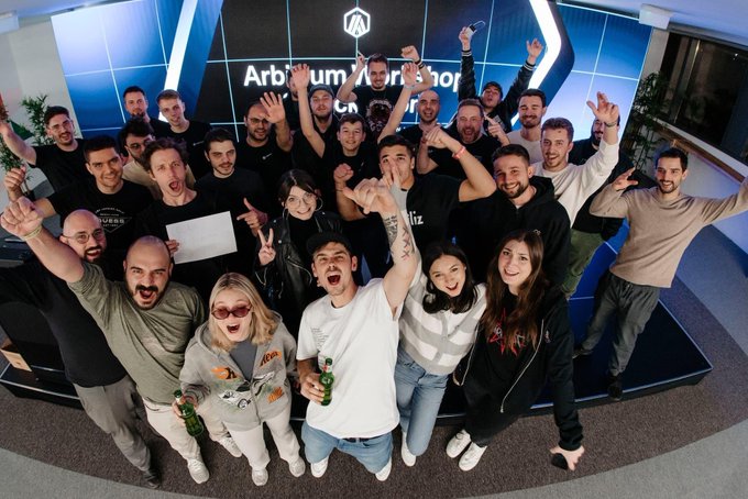 A large, energetic group of people celebrating together with raised arms and drinks in front of a screen displaying 'Arbitrum Workshop'.