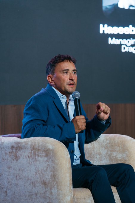 A man in a blue blazer sits in a cream-colored armchair, speaking into a microphone while gesturing with his hand, likely on stage at the Avalanche Summit as referenced in the post. Behind him, a screen displays partial text including "Haseeb" and "Managing," suggesting a speaker introduction. The image captures the engaging, community-focused atmosphere described in the post, with attendees sharing insights and reflecting on the event's growth.