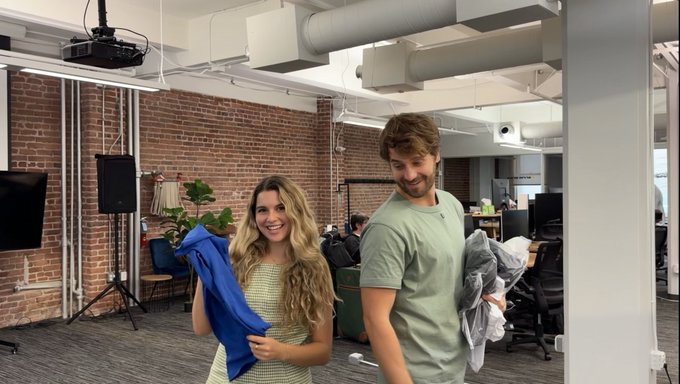 A woman with long blonde hair and a man in a green t-shirt smile in a modern office with exposed brick walls, the woman holding up a blue piece of clothing while the man holds a plastic bag—capturing a casual, friendly moment that complements the heartfelt caption thanking @danieldelophem for his support and guidance in her web3 journey.
