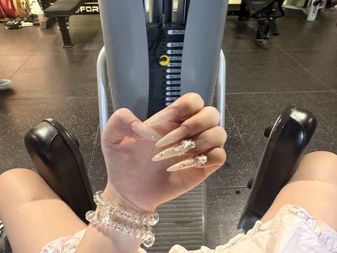 A close-up of a hand with long, glittery ombre nails adorned with 3D floral accents, paired with a clear beaded bracelet, posed against a gym leg press machine displaying "TECHNOGYM" and weight stack markings, while the person sits in light pink loungewear—capturing a morning gym moment paired with a crypto project announcement.