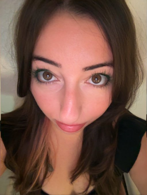 A close-up selfie of a young woman with long brown hair, striking brown eyes, and red lipstick looking directly into the camera.