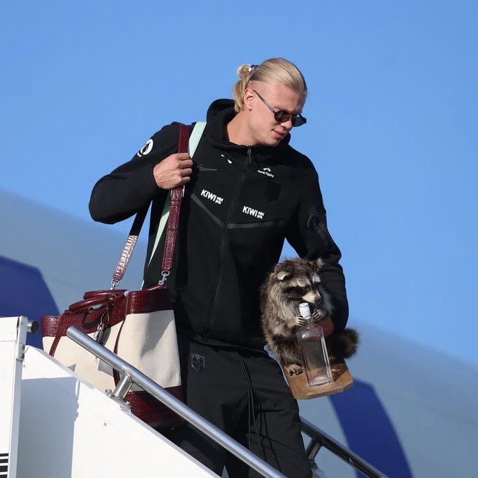 A blonde man wearing sunglasses and a black 'KIWI' jacket descends airplane stairs while carrying a raccoon that is drinking from a glass bottle.
