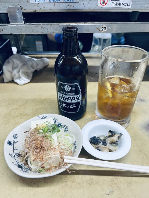 A Japanese meal at a counter featuring a bottle of Hoppy beer, a glass with a dark drink on ice, a plate of tofu topped with bonito flakes and scallions, and a small dish of pickled vegetables.