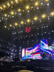 A concert venue interior featuring elaborate stage lighting rigs, large LED screens displaying colorful visuals, and an illuminated circular logo above the stage.