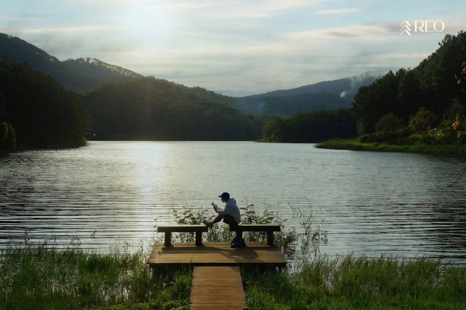 A person wearing a hat sits on a wooden bench at the edge of a tranquil lake, reading a book, with the logo 'REO' in the corner.