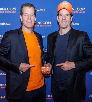 Two men pose together at a Bitcoin 2025 event; one wears a black jacket over an orange shirt, and the other wears an orange cap with a Bitcoin logo, a dark jacket, and holds a small 'Bitcoin' sign.
