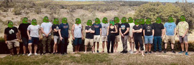 A group photo of people standing outdoors in a line with their faces covered by green Pepe frog masks, wearing casual clothing against a backdrop of trees and hills.