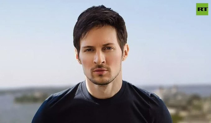 The image shows a handsome young man with dark hair and a light beard, wearing a black t-shirt, posing outdoors with a cityscape in the background. The "RT" logo is visible in the top right corner, and the post describes him as a TON crypto developer who has been arrested after being internationally wanted.