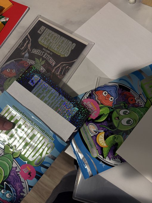A hand holding a holographic 'Wall Street Memes' collector's comic pack featuring cartoon characters, alongside a 'Welcome to Shells Kitchen' DVD or print.