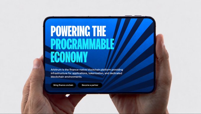 A pair of hands holds a smartphone displaying the Arbitrum homepage with the text "POWERING THE PROGRAMMABLE ECONOMY" and a description noting Arbitrum as "the finance-native blockchain platform providing infrastructure for applications, tokenization, and dedicated blockchain environments," alongside "Bring finance onchain" and "Become a partner" buttons—visually representing the post's quip about the "new iPhone, but programmable" by showcasing Arbitrum as a programmable financial device for the onchain economy.