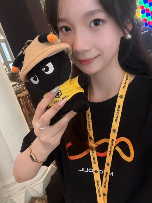 A young woman wearing a black t-shirt with an infinity logo holds a black plush mascot wearing a yellow BNB (Binance) hat; she wears a lanyard reading 'BNB CHAIN'.