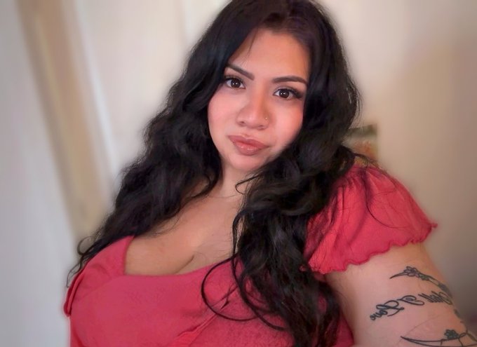 A woman with long dark wavy hair, wearing a red top, poses for the camera while a tattoo with partially visible cursive text reading "R" appears on her arm. Her confident and steady gaze complements the post's message about perseverance and staying focused on long-term goals through market downturns.
