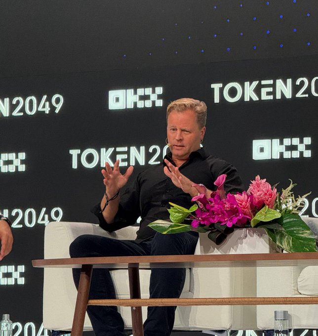 A man in a black shirt sits on a white couch on a stage at 'TOKEN2049', gesturing with his hands while speaking next to a wooden table holding a vibrant floral arrangement.