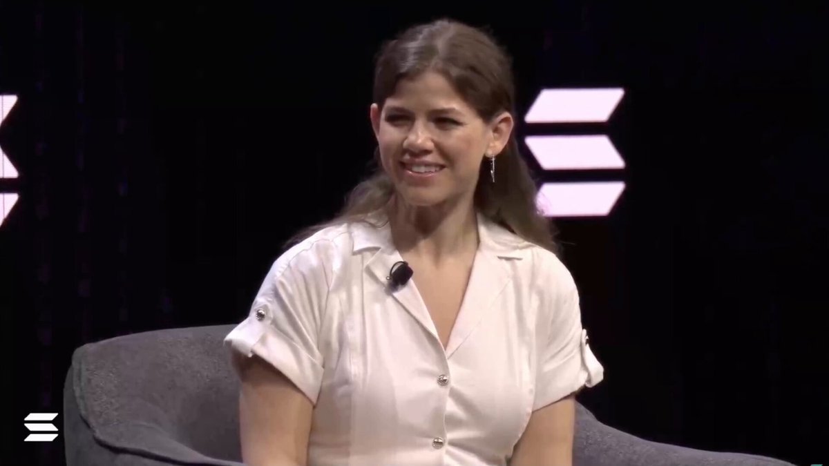 The image shows a woman wearing a white dress sitting in a chair with her hands at her sides. She appears to be smiling as she looks directly into the camera. The background of the image features a black curtain, which contrasts with the white dress and adds depth to the scene. There are also some text elements present in the image that read "@SoFi picked Solana" and "@Solana is the right chain to use for payments because of the cost, settlement speed and ultimately the throughput".