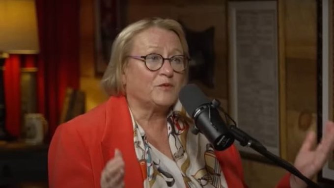 A woman with short blonde hair, glasses, and a red blazer speaking into a microphone during what appears to be a podcast recording.