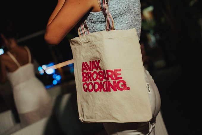 A close-up shot shows a person walking at night carrying a beige tote bag with bold red text reading 'AVAX BROS ARE COOKING.', likely referencing the Avalanche (AVAX) cryptocurrency community.