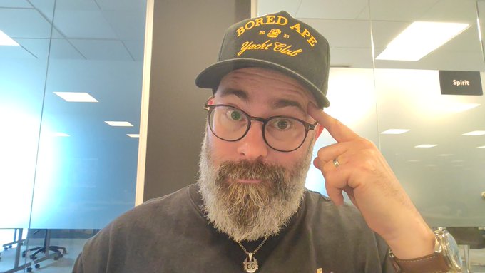 The image features an office setting with a man wearing glasses, a hat, and a beard standing next to a desk. The man has his hand up to his head, suggesting he might be thinking or pondering something. There are also two clocks in the background of the image, one on top of another. The post includes text that reads "Office Policy 101: Ape Shall reward Ape A Bored Ape is a way of life". This message seems to be related to the man's situation and could possibly be an office joke or a reference to a popular saying about boredom.