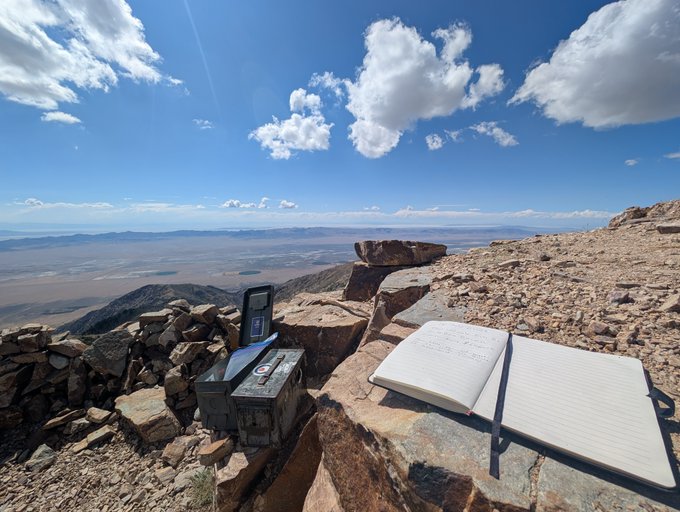 A scenic mountain summit with a journal, pen, and GPS device resting on rocks overlooking a vast desert landscape under a blue sky with scattered clouds.