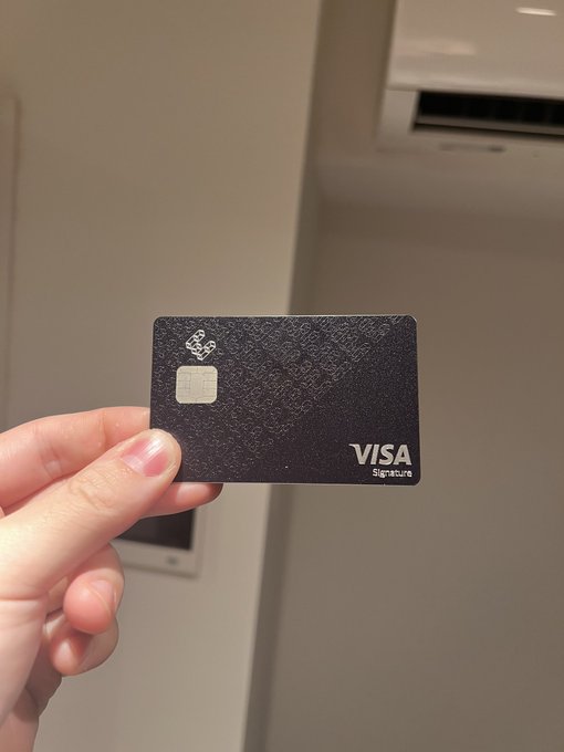 A hand holds up a black Visa Signature credit card with a chip and decorative pattern, photographed in front of a wall-mounted air conditioning unit.