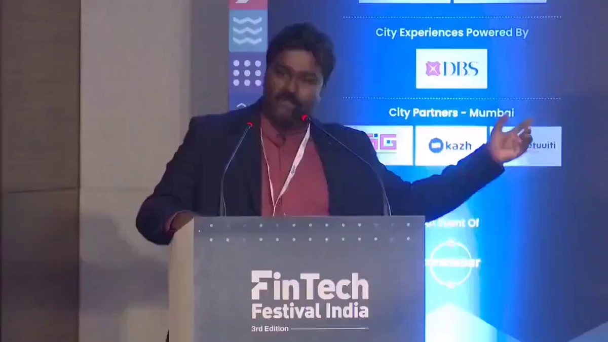 The image shows a man standing behind a podium at an event, giving a speech or presentation. The man appears to be wearing a suit and has his arms outstretched as if he's making a gesture during his talk. In front of him is a sign that says "City Experiences powered by DBS", indicating the sponsor of the event. The background features a blue screen displaying various logos, including one for Ripple, which suggests that this might be an international conference or gathering related to finance and technology.