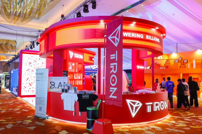 A red and orange illuminated TRON exhibition booth displaying merchandise and the phrase 'EMPOWERING BILLIONS' at an indoor event.