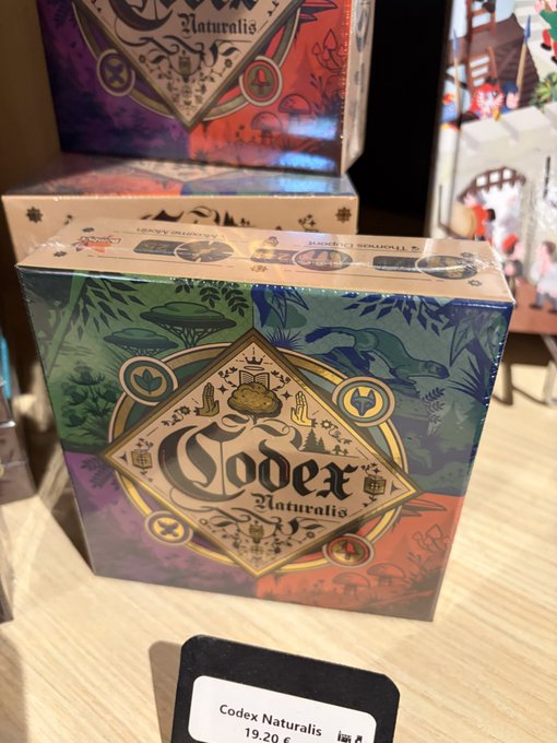 The image shows a box of the board game "Codex Naturalis" priced at 19.20€, aligning with the post mentioning it being available for 20€. The game features decorative artwork with mushrooms and wildlife on its cover, displayed on a shelf alongside other board games.