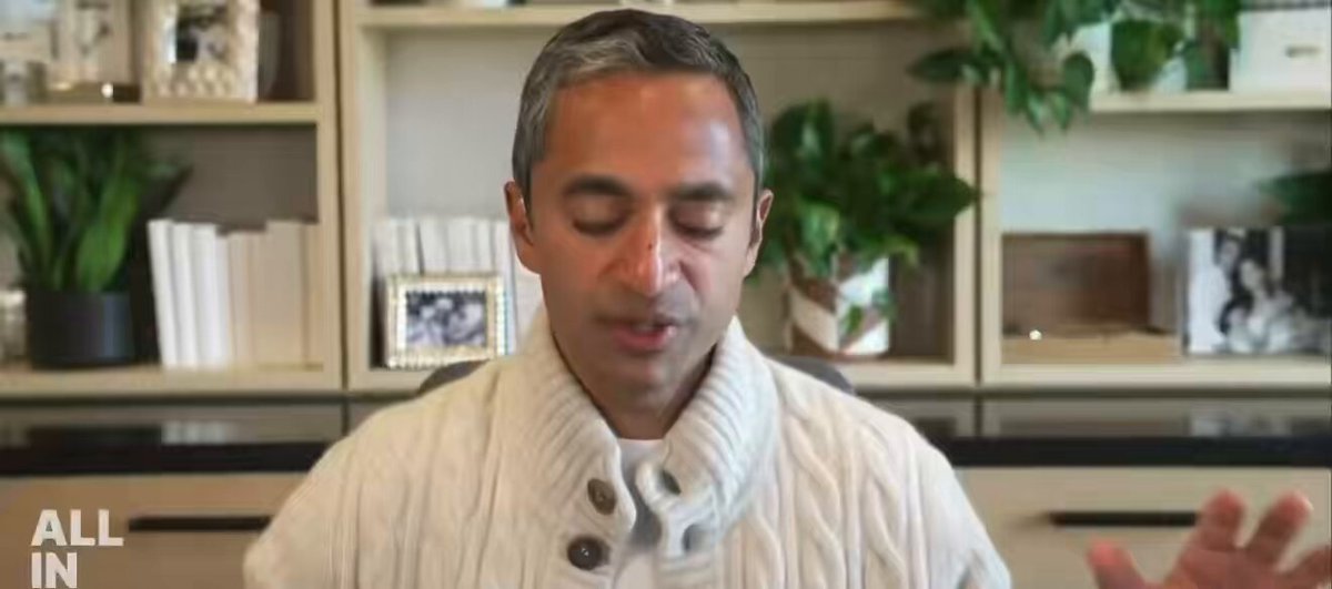 The image features a man wearing a white sweater, sitting at a desk with his hands outstretched as if he's giving a speech or presentation. The background includes several potted plants placed around the room, adding a touch of greenery to the scene. There are also books scattered on the table and shelves behind him, suggesting that this might be an office setting where the man is discussing AI-related topics with others.