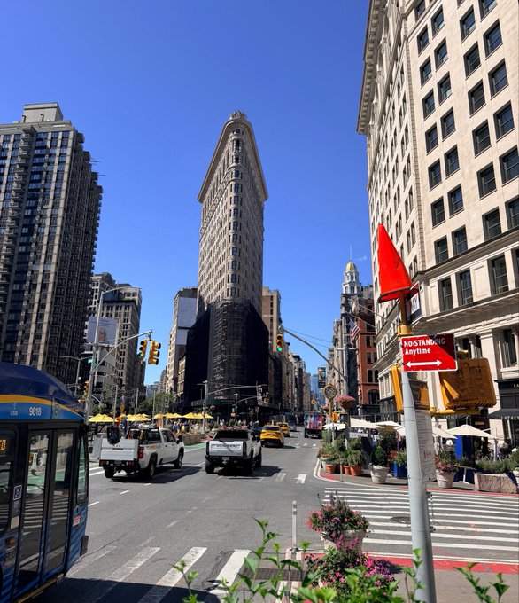 The image shows a busy city street with tall buildings, cars driving down the road, and people walking along the sidewalks. There is also an orange traffic sign that reads "No Star St." near the center of the scene. The photo appears to be taken from across the street at a bus stop or intersection, capturing the bustling urban environment.