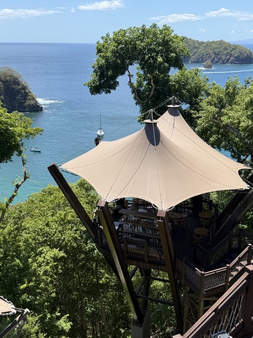 A scenic photograph shows a tropical treetop structure with a peaked canopy roof overlooking a blue ocean with rocky islands, featuring a bar area inside the elevated wooden platform.