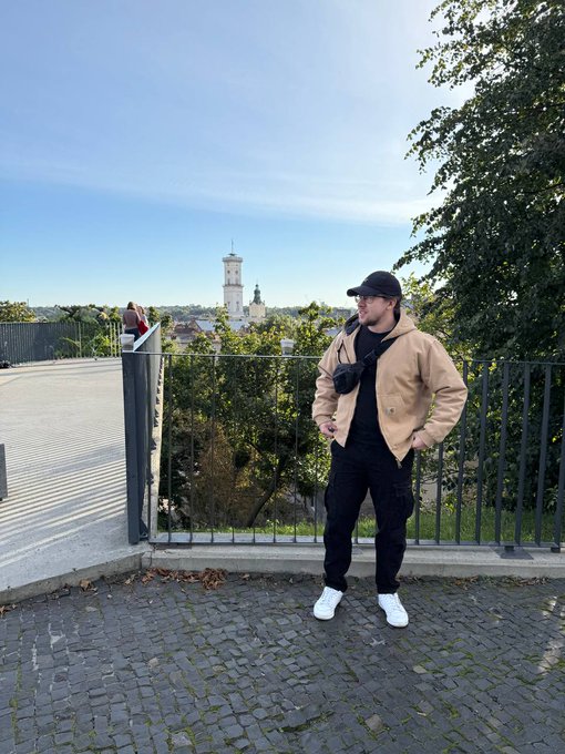 A man wearing a tan jacket, black cap, and glasses stands on a cobblestone overlook with a metal railing, with a town featuring a church tower visible in the background.