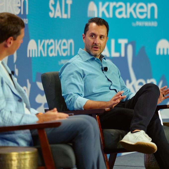 Two men sit on stage at the SALT Conference, with the backdrop displaying "Kraken" and "SALT" logos as the speaker in a light blue shirt gestures while conversing, consistent with the post's mention of the Wyoming conference gathering finance, policy, and digital assets industry leaders.