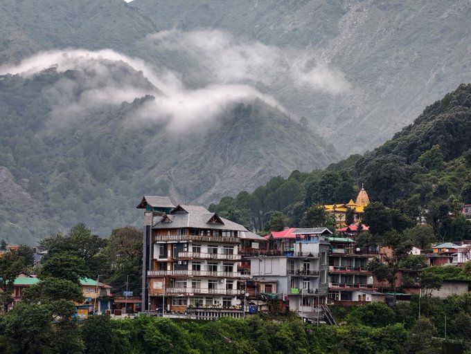 Mist-covered mountains rise behind a hillside town featuring multi-story buildings and a temple with a golden dome nestled among trees.