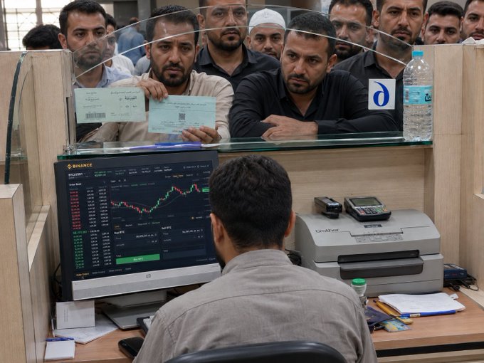 A group of men crowd around a counter at what appears to be a currency exchange office, watching as an employee focuses on a Binance chart displayed on his monitor showing BTC and ETH trading data, while a Brother printer sits beside him—capturing the irony of calmly monitoring crypto charts amidst an urgent transaction.