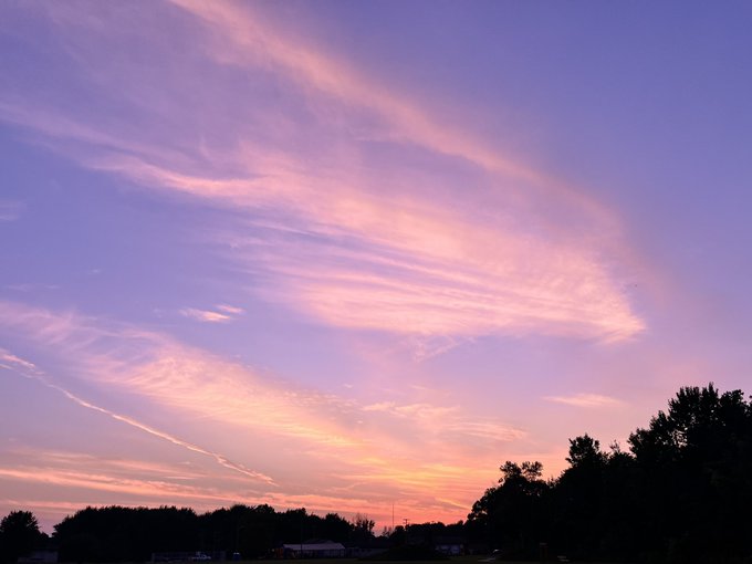A serene sunset sky with wispy pink and orange clouds against a lavender-blue backdrop, with silhouetted trees visible along the bottom.