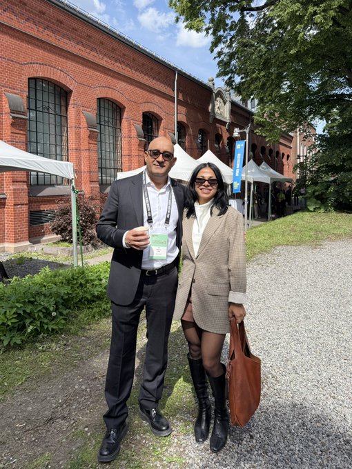 A man in a suit and a woman in a blazer and thigh-high boots pose together on a gravel path in front of a brick building.