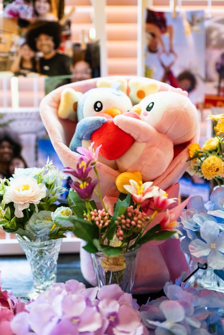A plush toy bouquet featuring a penguin character holding a heart, accompanied by colorful flowers including daisies, lilies, and hydrangeas in vases, with the background showing promotional posters of people—matching the "Pengu bouquet" mentioned in the post.