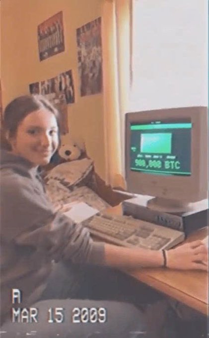 A young woman smiles beside an old CRT monitor displaying "900,000 BTC," dated "MAR 15 2009," playfully parodying the "looking for my lover" meme by joking that she'd trade 900,000 Bitcoin (now worth billions) just to have her partner appear in the photo beside her.