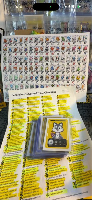 Photo showing a 'VeeFriends Series 2 TCG Checklist' document beneath a stack of VeeFriends trading cards, with a sheet of character sticker icons on a wall in the background.