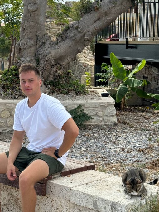 A young man in a white t-shirt and green shorts sitting on a stone bench outdoors next to a tree, with a gray and white cat resting on the ground beside him in what appears to be a Mediterranean setting.