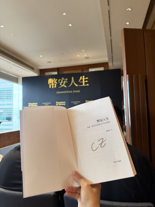 A person holding open a book titled 'Chuangeng Zhao' (币安人生) showing a signature, with a backdrop displaying 'Freedom of Money' text.