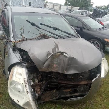 A photograph of a silver car with significant front-end damage, including a crumpled hood, broken headlight, and exposed engine components, appearing to be in a salvage yard with other vehicles in the background.