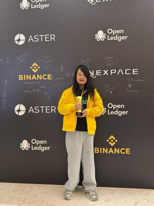 A person in a yellow jacket holds a trophy in front of a step-and-repeat banner featuring logos for Binance, Aster, Open Ledger, and NEXPACE, surrounded by handwritten signatures. The image connects to the post by showing the winner proudly displaying the "Binance's Annual Popularity King" trophy mentioned in the story.
