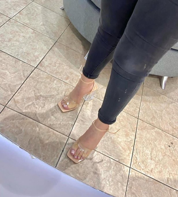 A person's legs wearing dark jeans and tan/beige open-toe high-heeled sandals with ankle straps, standing on a tiled floor.