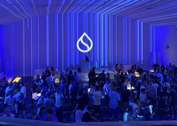 Crowd of people at an event bathed in blue lighting with a large water droplet logo on the wall, resembling a Sui or similar crypto event.